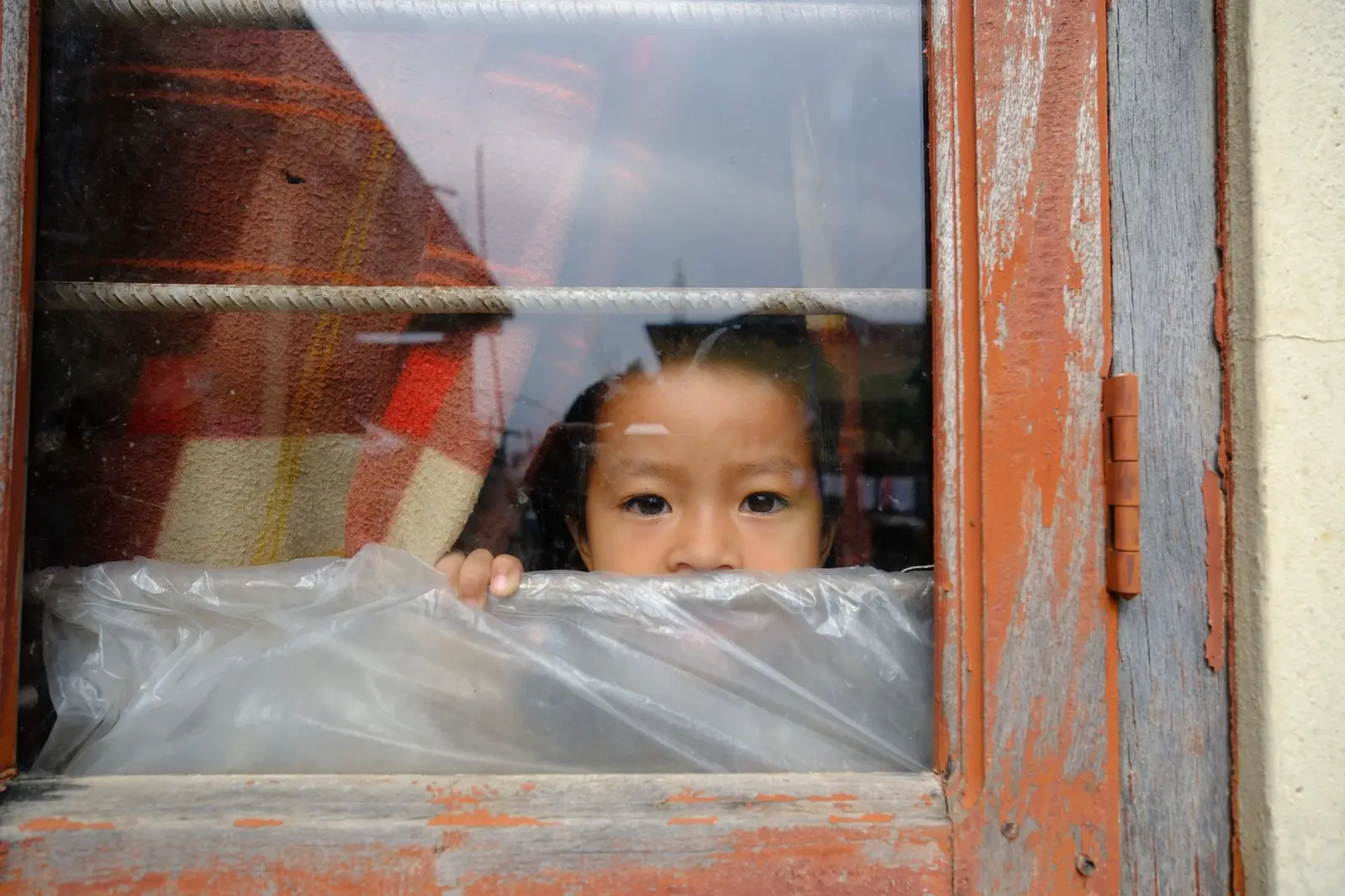 A young child stands at a camp window, face pressed near the glass and a sheet of plastic, looking directly out through a weathered orange door frame.