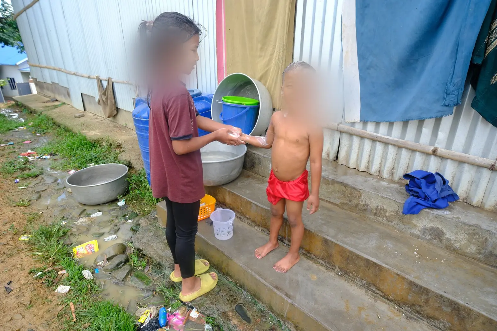 A small child, his face blurred for protection, is washed on a concrete step an arm’s length from a heap of wet rubbish at the edge of the camp.
