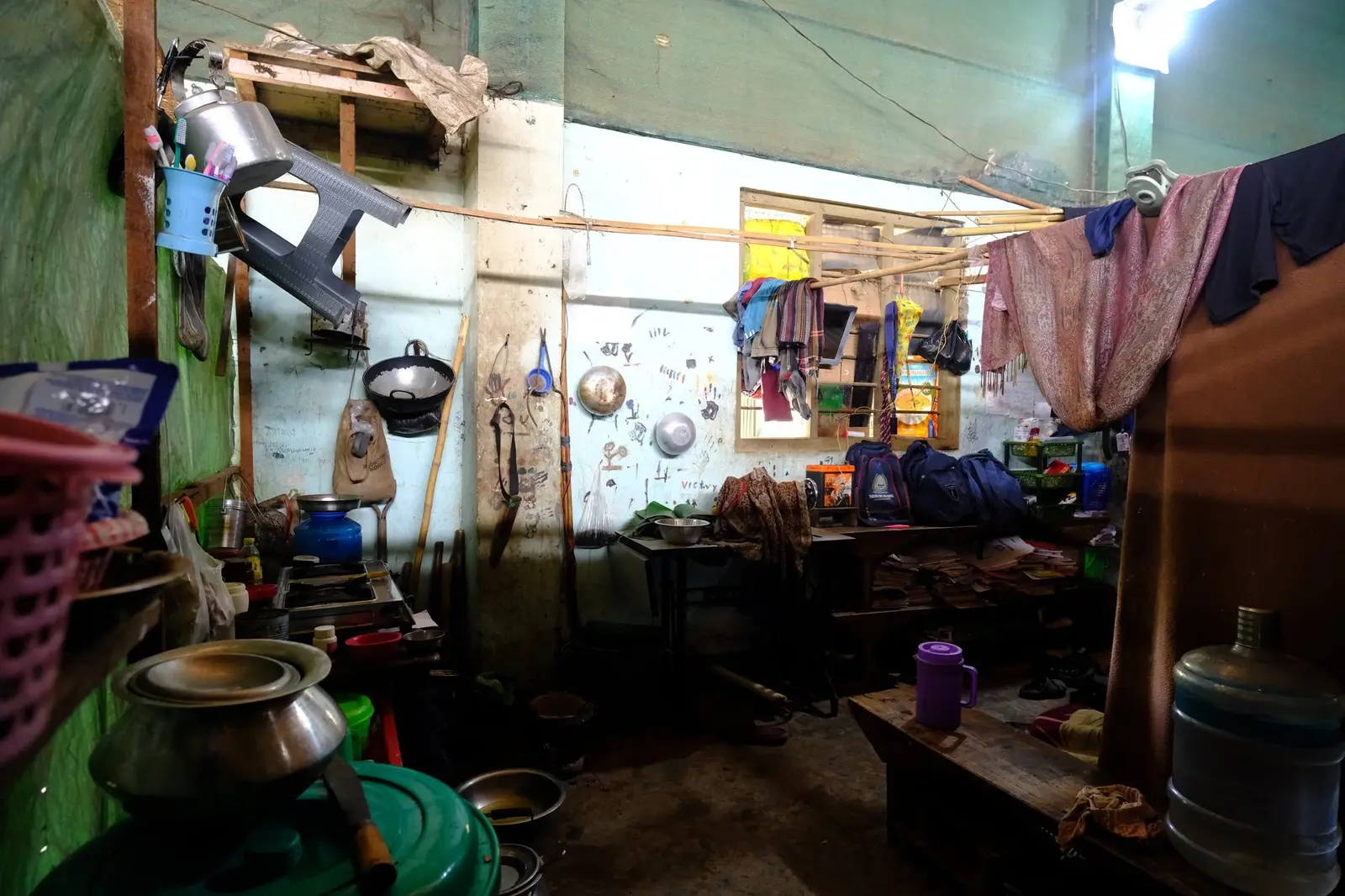 A corner of a camp room used as a kitchen — a small stove and a few pots set on the floor, the family's school bags hung on the wall, and a child's handprints pressed into the paint above them.