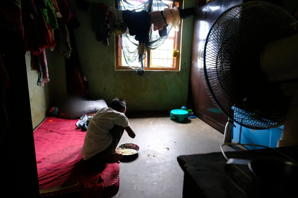A child, face blurred for protection, sits alone on the floor of a dim room eating from a single steel plate, a pedestal fan and a scatter of toys around them.