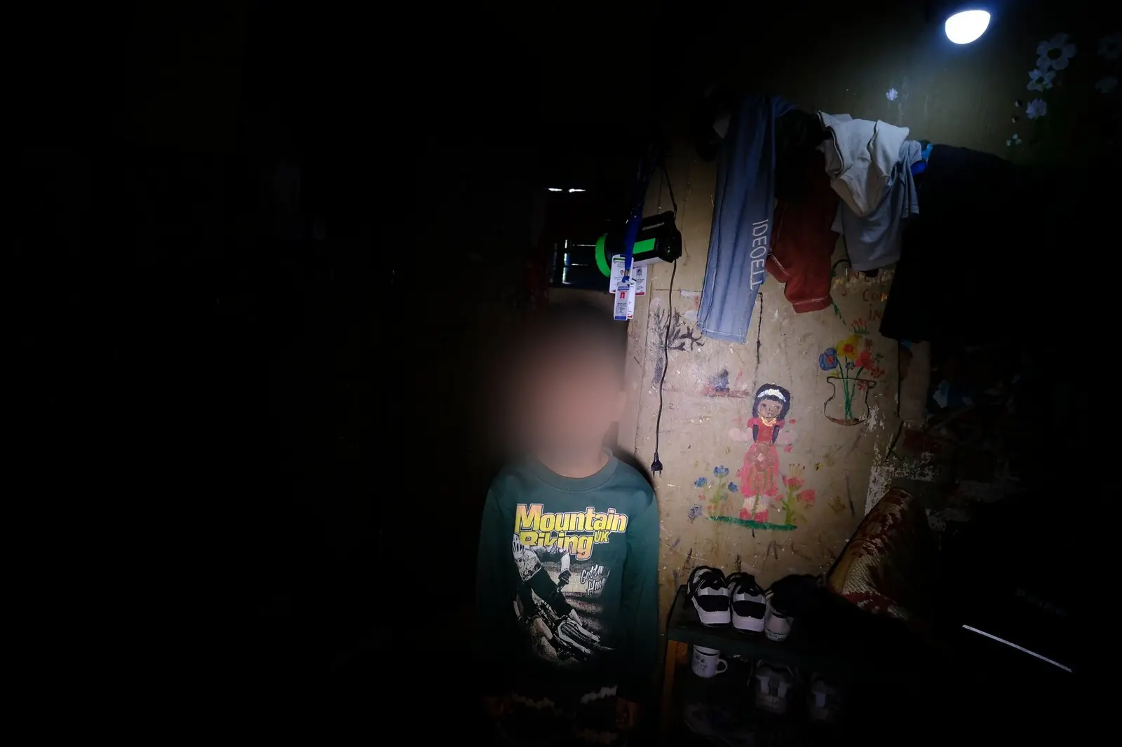 A boy, his face blurred for protection, sits in a dim camp room; on the wall behind him a child has painted a girl standing among flowers.