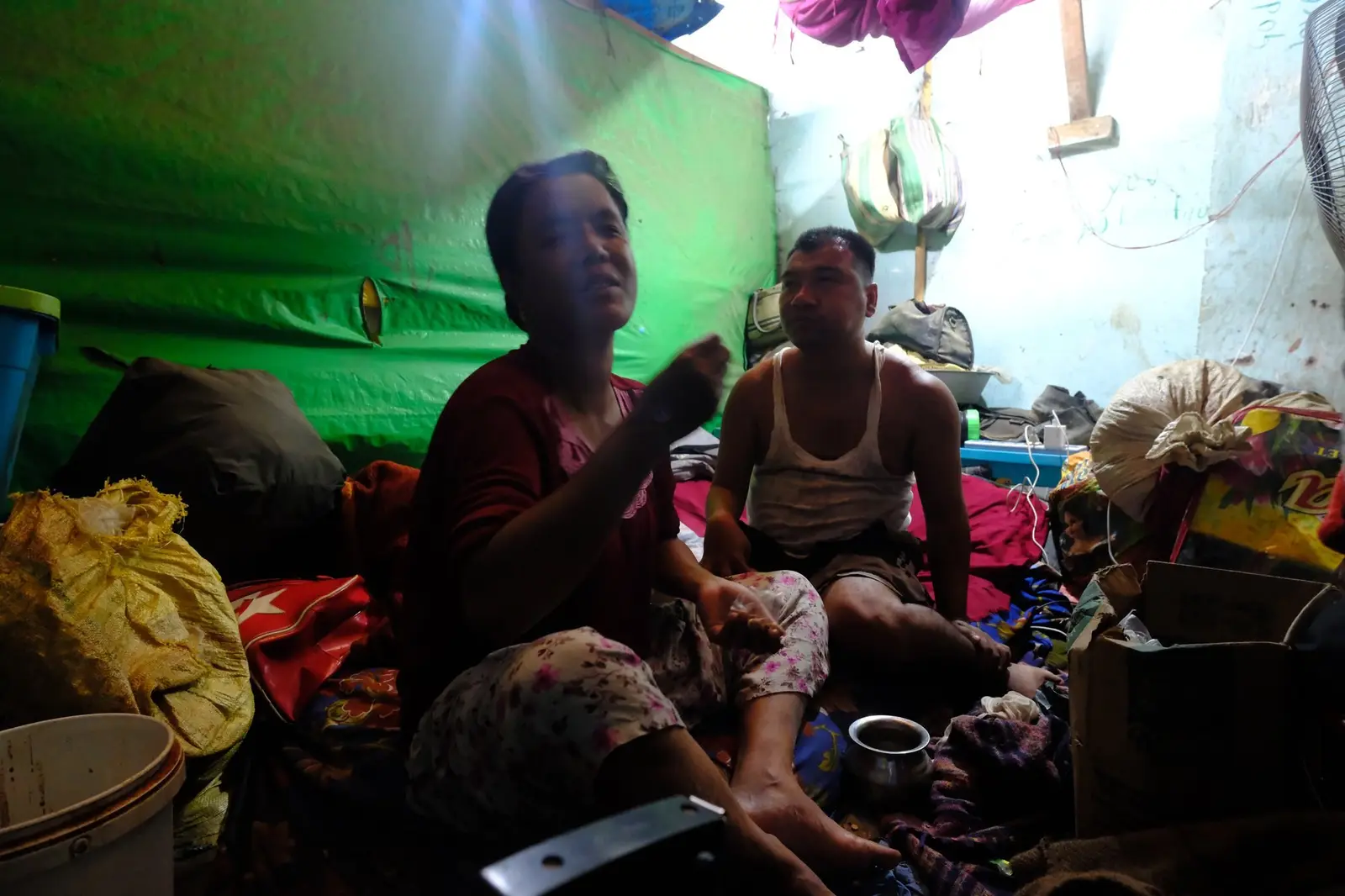 A couple from Moreh sit among everything they salvaged — bundles, trunks, pots and bedding stacked floor to ceiling and balanced wall to wall — pressed into a single camp room smaller than a bed.