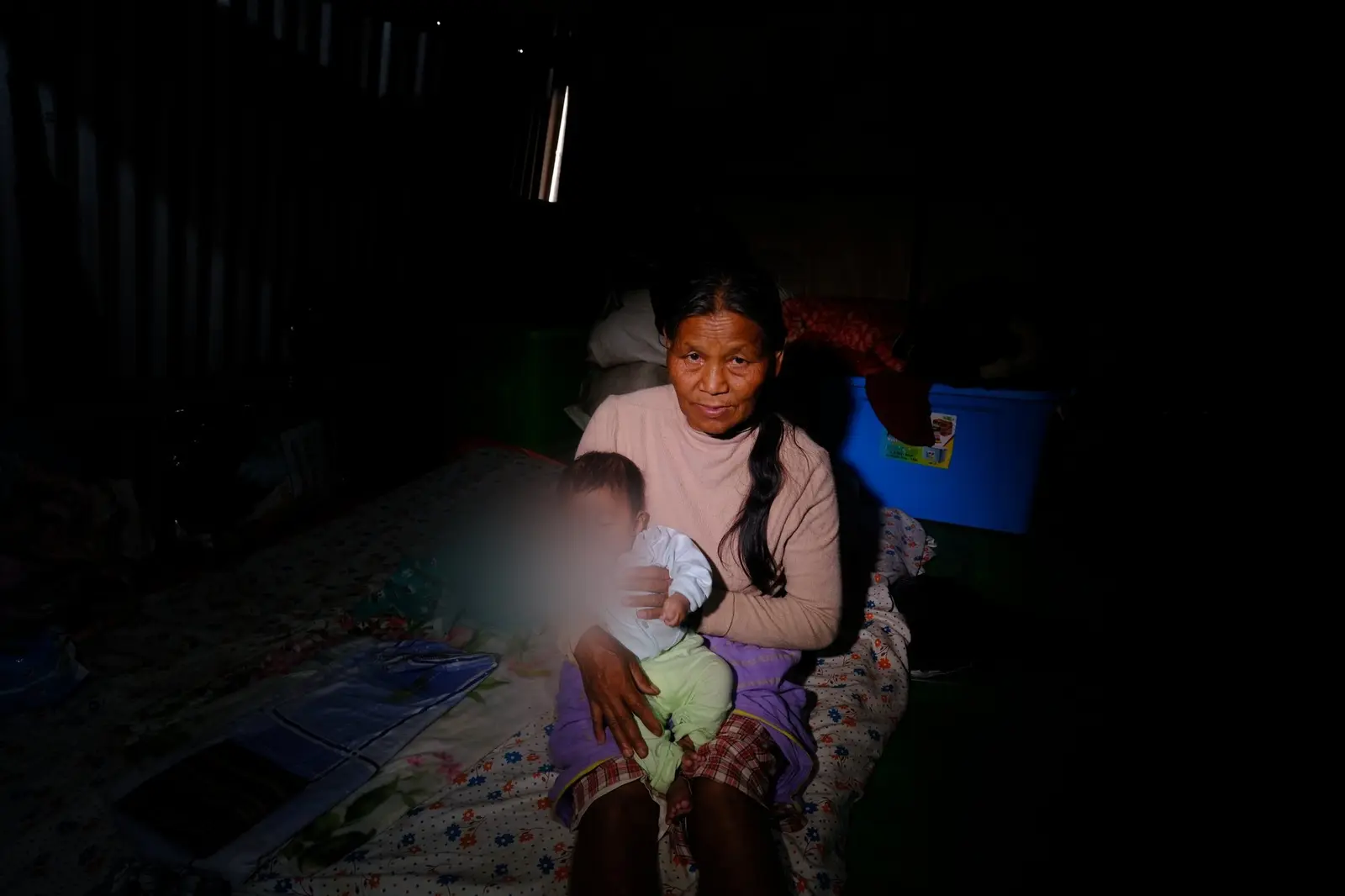 An elderly grandmother cradles a sleeping infant, the child's face turned away, in a dim and almost unlit camp room — the oldest and the youngest of one displaced family.
