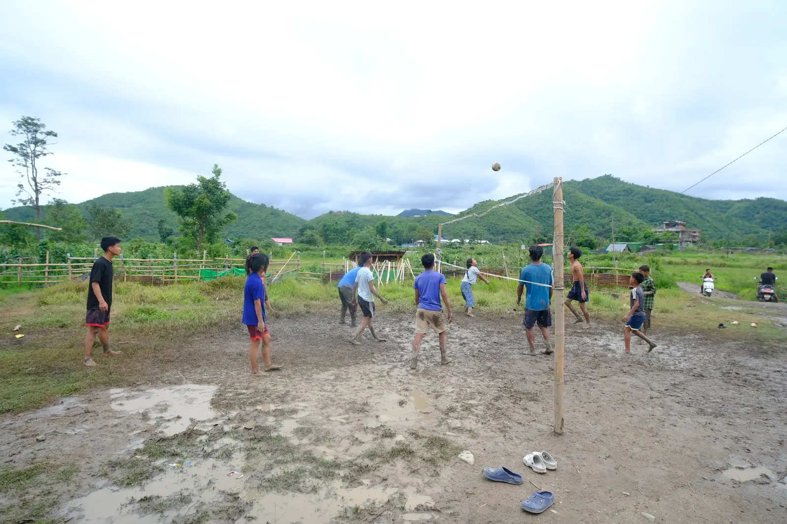 A volleyball court improvised from a net of string slung between two bamboo poles, pitched over a field of churned mud, with the camp shelters crowding the far edge.