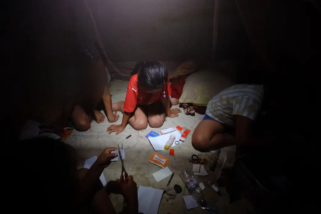 Children's hands, their faces blurred for protection, sorting medicine boxes and a pair of scissors into a game beneath a mosquito net, lit by a single bulb.