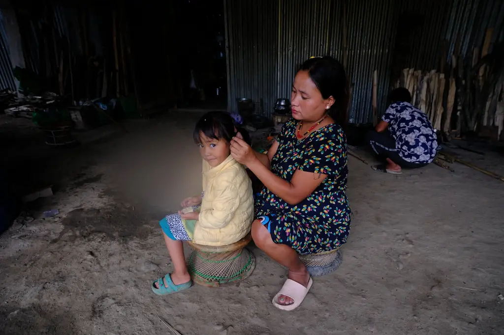 A mother sits on the bare earth braiding her daughter's hair, the child's face blurred for protection.