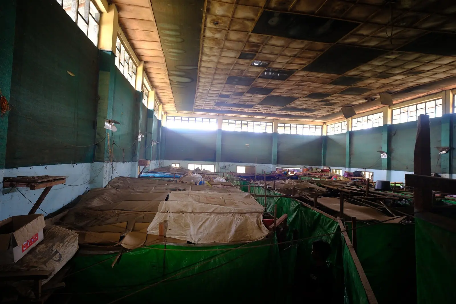 One long hall partitioned by hanging tarpaulin sheets and string into a grid of small household squares, the row of makeshift rooms receding into the dim.