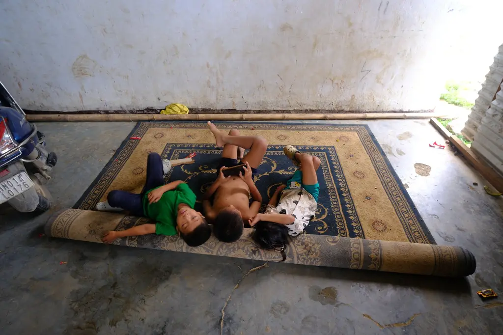 Three children, their faces blurred for protection, crouch over a single phone on a half-rolled carpet in a camp corridor.