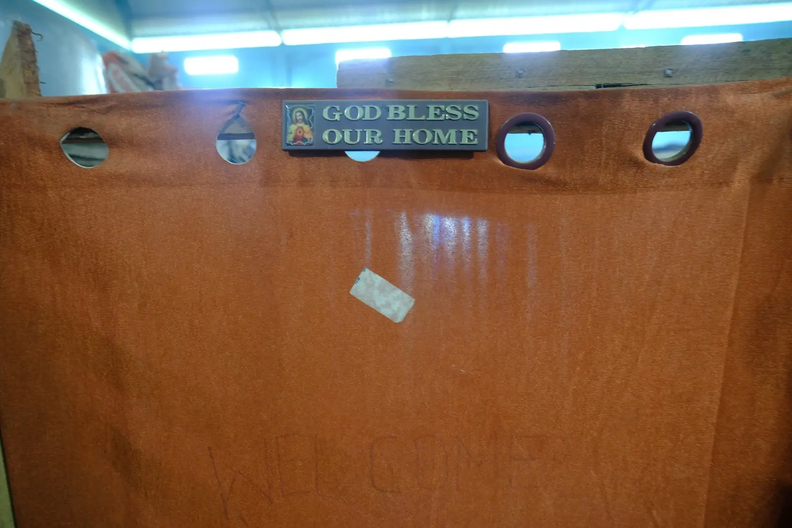A partition wall of blue plastic sheeting inside a camp, a small printed plaque fixed to it that still reads "God bless our home."