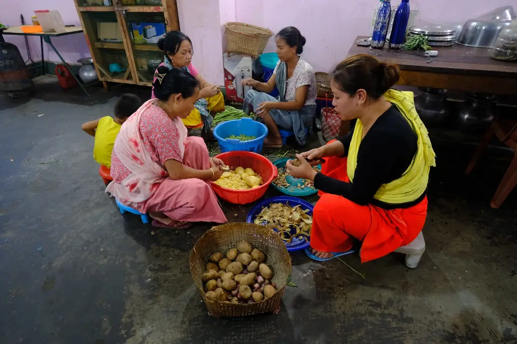 Women sit together on a swept floor in a Thangmeiband camp kitchen, peeling vegetables into steel bowls, the room lit and tended with pots stacked along the wall.