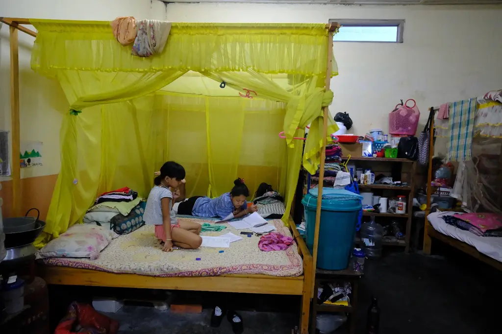 Four young siblings, the smallest faces blurred for protection, sit close together on the bed of their tarpaulin-walled partition, folded clothes and a tidy shelf behind them.