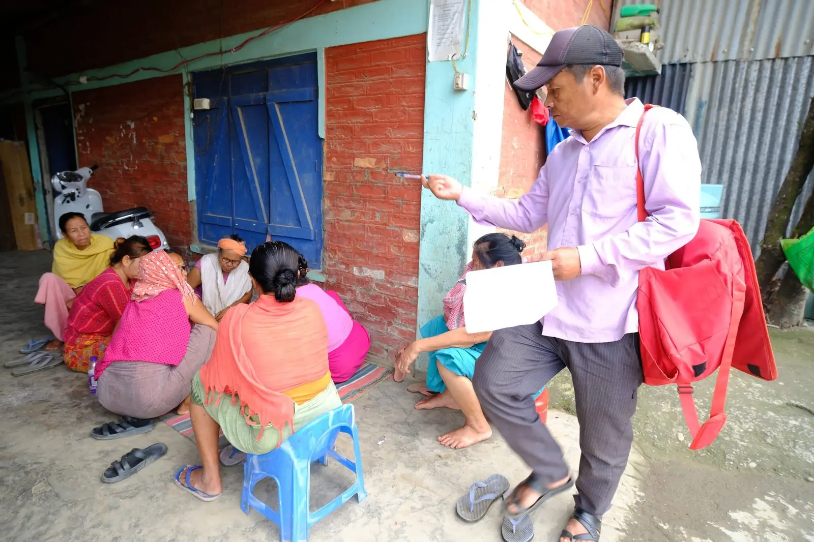 A camp outreach worker in a cap and red shoulder bag stands holding a sheet of paper, speaking to a group of women seated on stools and on the ground outside a brick camp building.