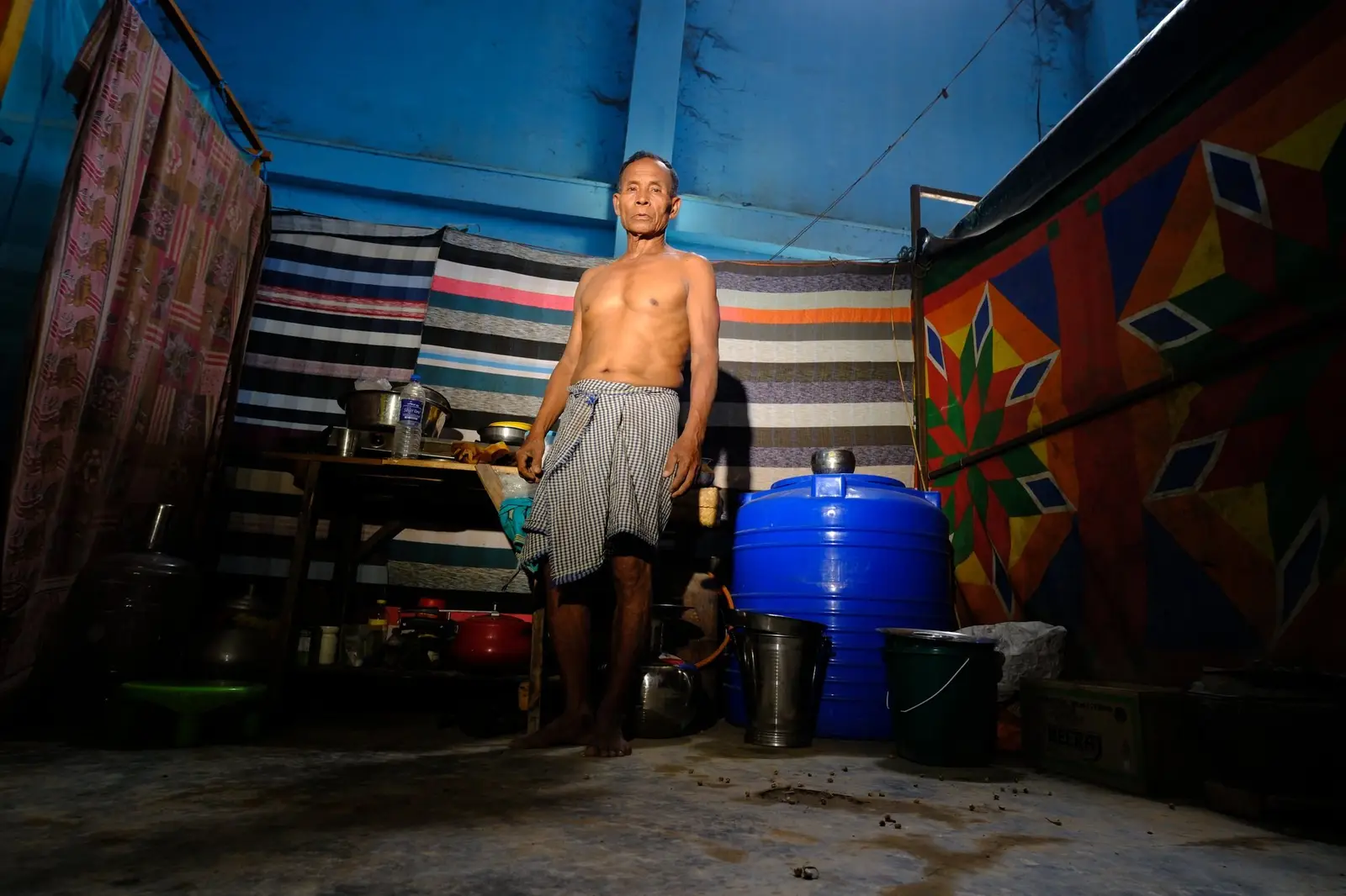 A man stands barefoot in his single room in Churachandpur, a blue water tank, drums and buckets ranged along the wall beside a striped cloth partition.