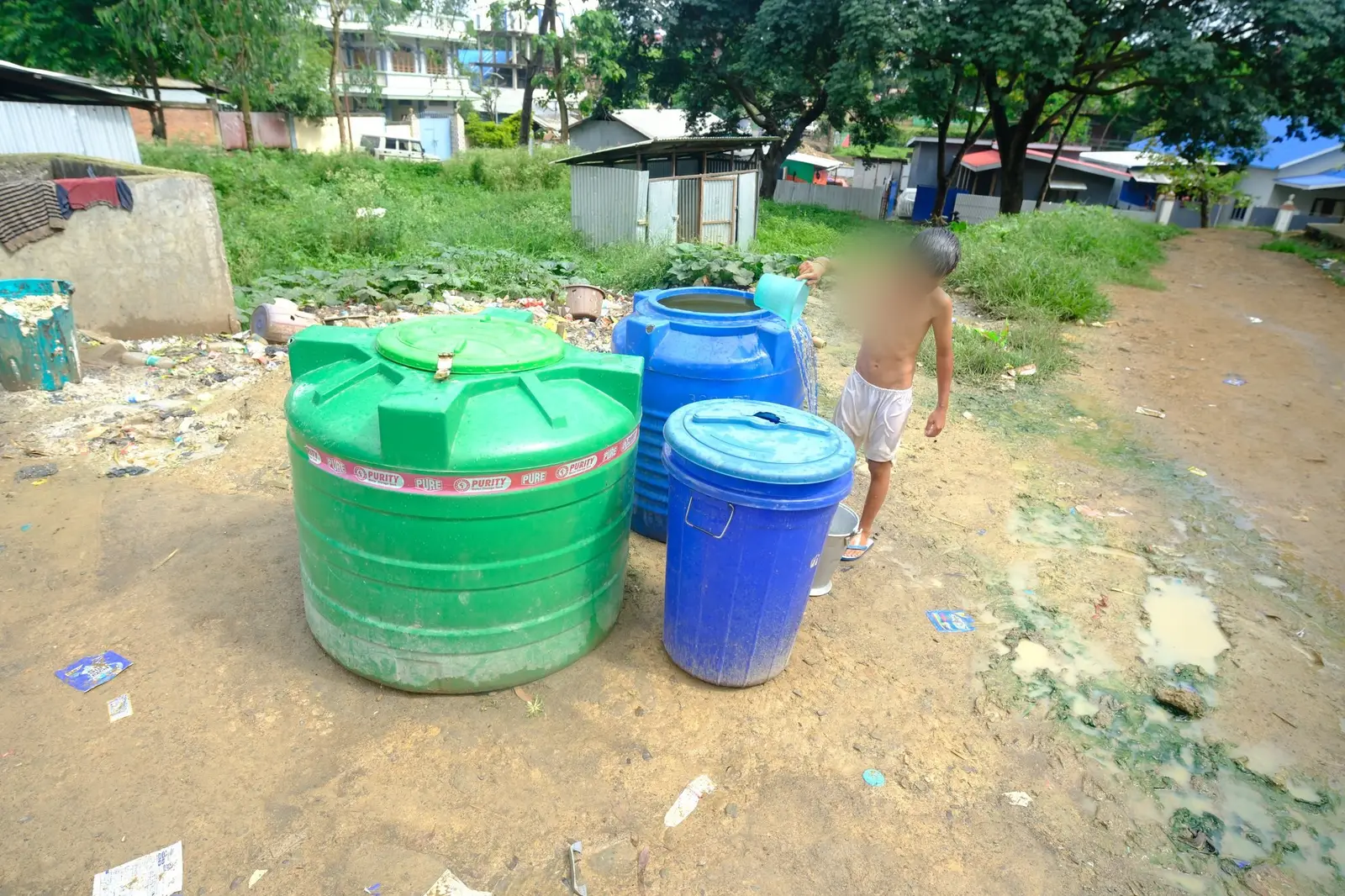 A boy, his face blurred for protection, bathes from green and blue household water drums on bare dirt at the edge of an open dump.