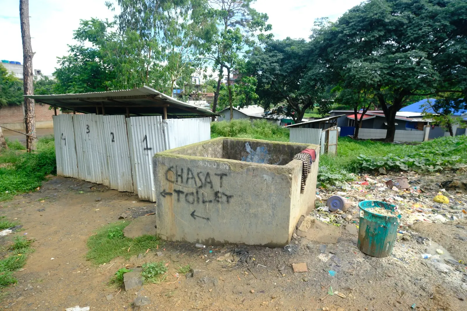 Four corrugated-tin toilet stalls and a water tank stand in a row on bare ground at the lip of an open dump, their doors weathered and hanging.